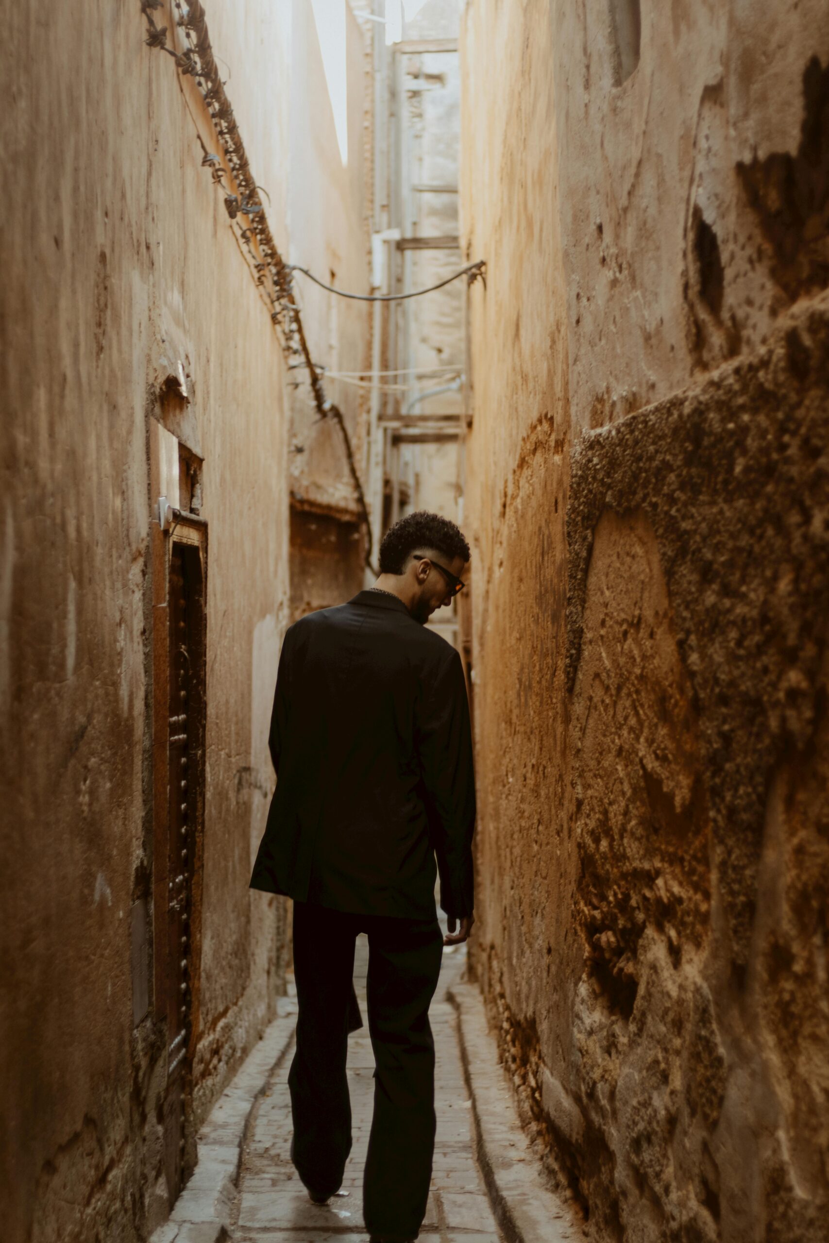 A man in a black suit walks down a narrow, historic alleyway with textured walls.